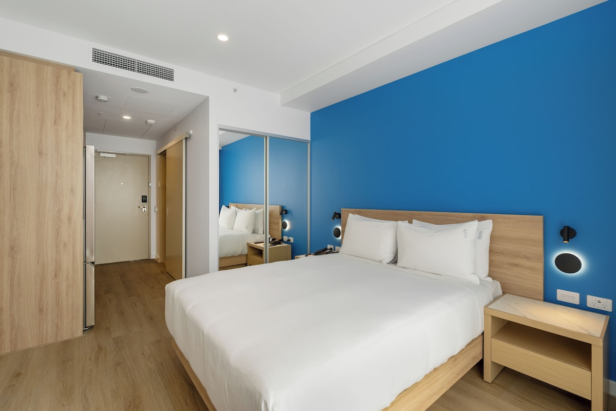 A modern hotel room with a large bed, bedside tables, a mirror, and a blue accent wall.