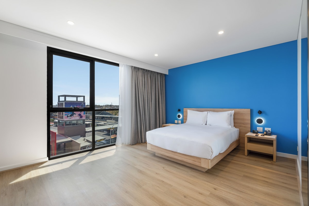A modern hotel room with a large bed, bedside tables, and a view through a floor-to-ceiling window.