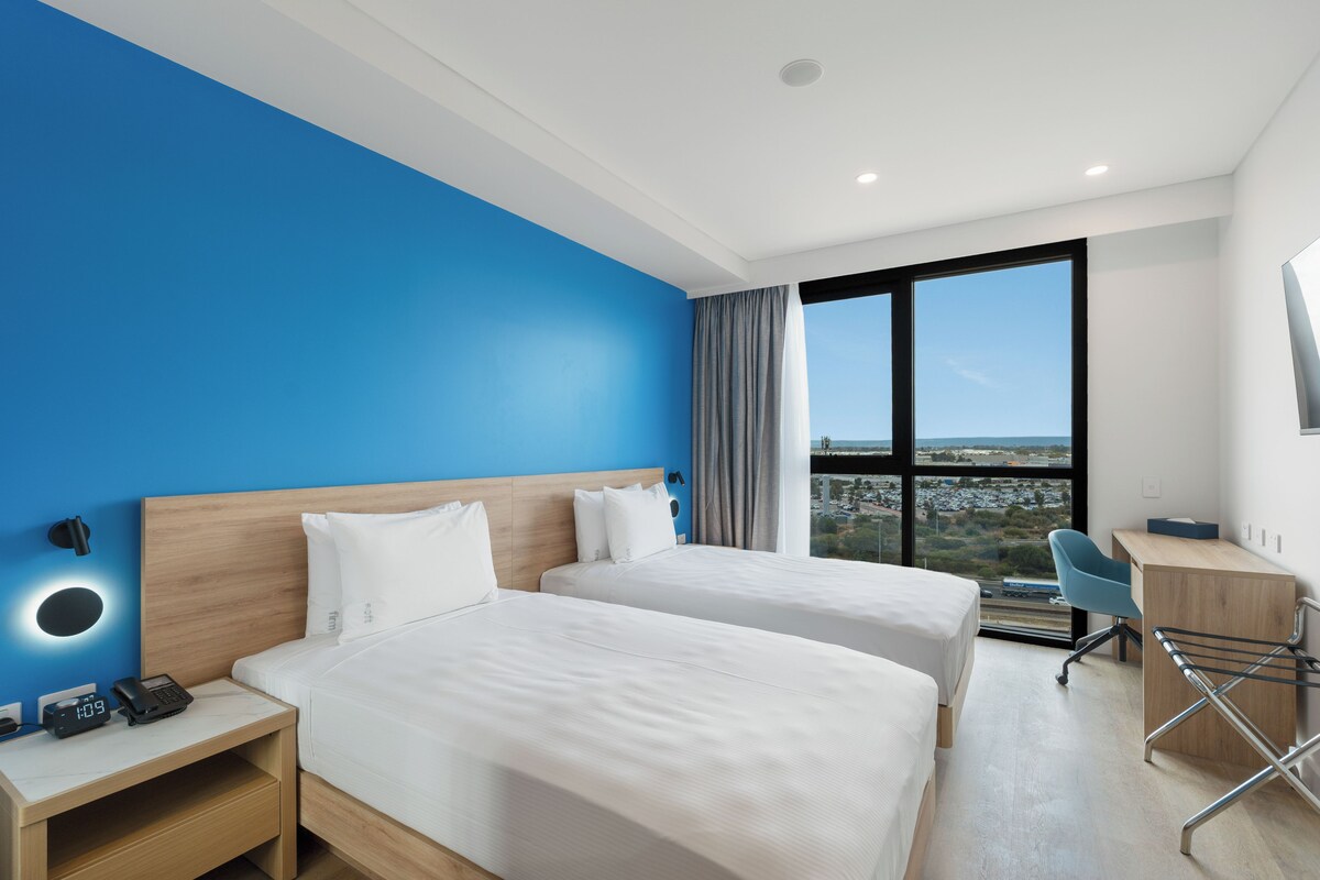 A hotel room with two beds, a desk, a chair, and a large window with a city view.