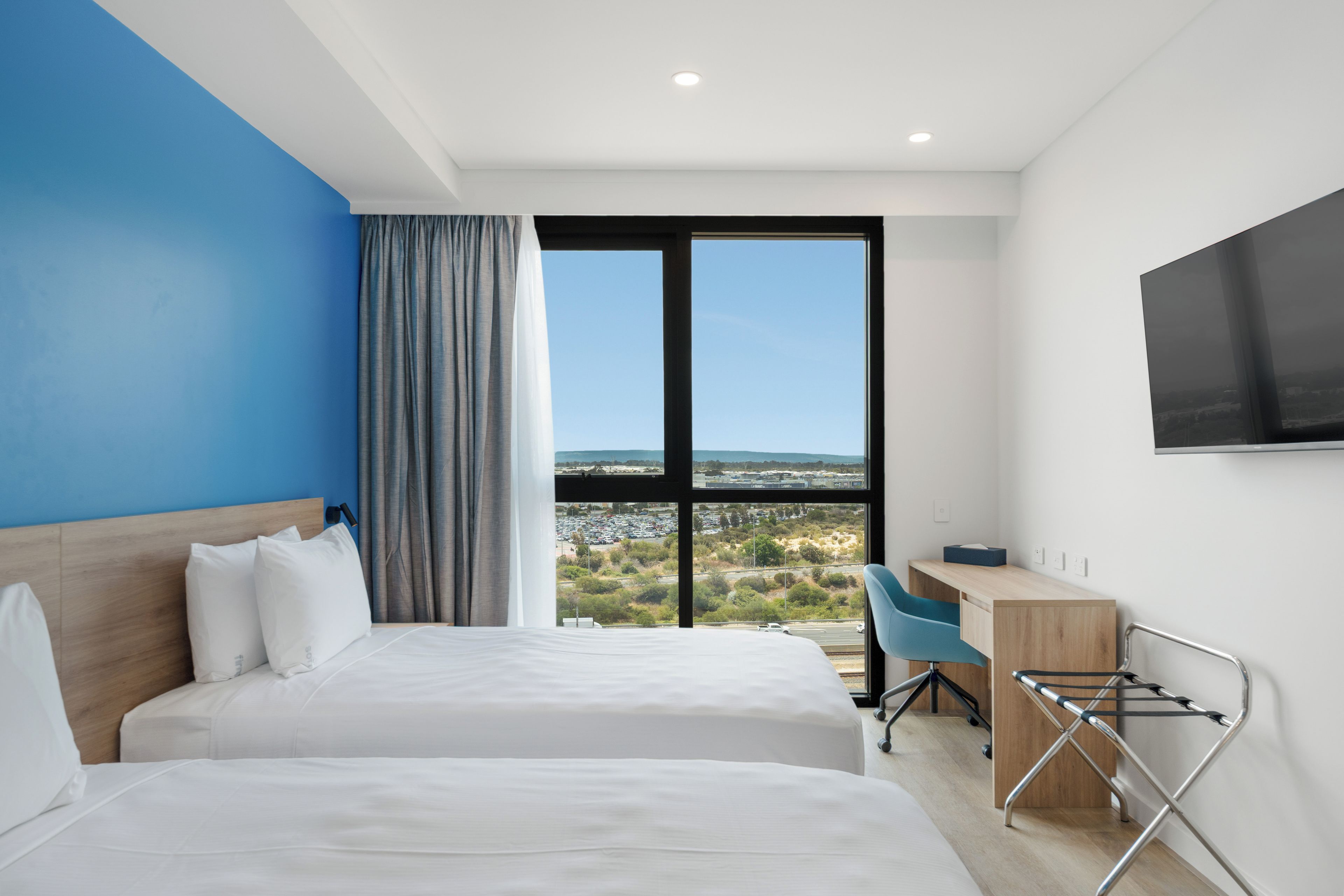 A hotel room with two beds, a desk, a chair, a TV, and a large window with a view of a parking lot and greenery.