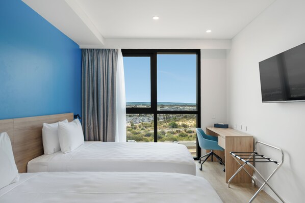 A hotel room with two beds, a desk, a chair, a TV, and a large window with a view of a parking lot and greenery.