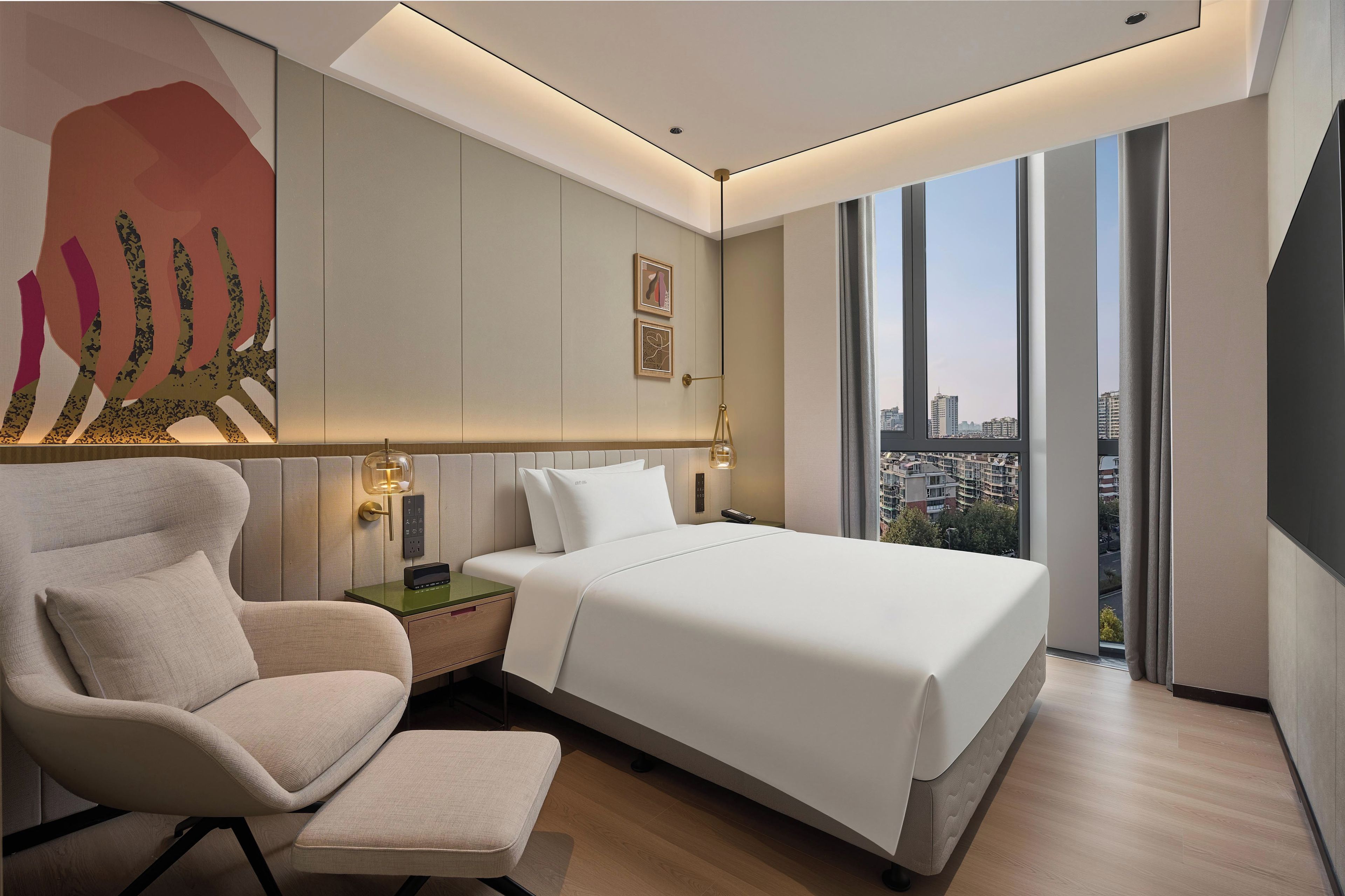 A modern hotel room with a large bed, a sitting area with a chair, a nightstand, and a wall-mounted lamp.
