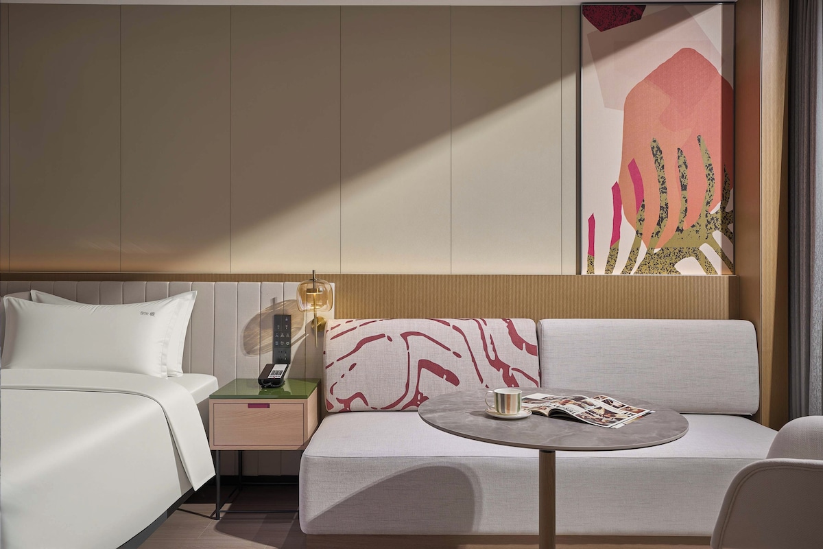 Habitació d'hotel amb un llit, una tauleta de nit, una làmpada, un sofà i una taula petita amb una revista.
