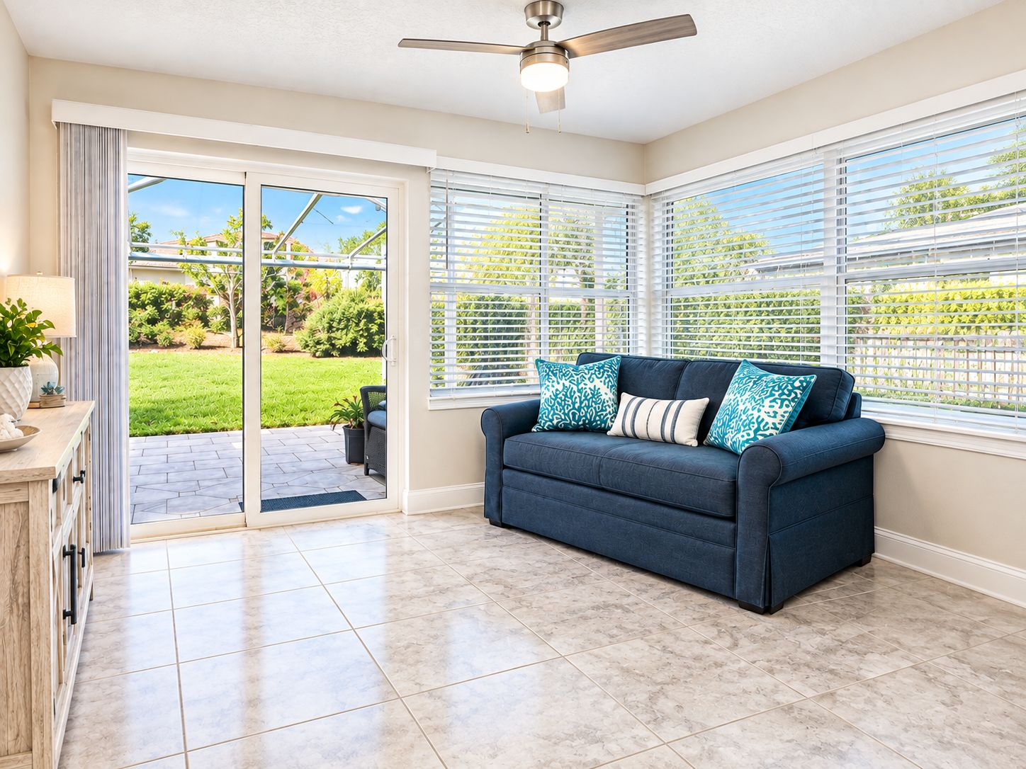 A room with a dark blue sofa, a ceiling fan, and a view of a green garden through large sliding glass doors.