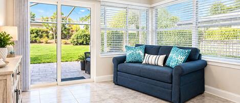 A room with a dark blue sofa, a ceiling fan, and a view of a green garden through large sliding glass doors.