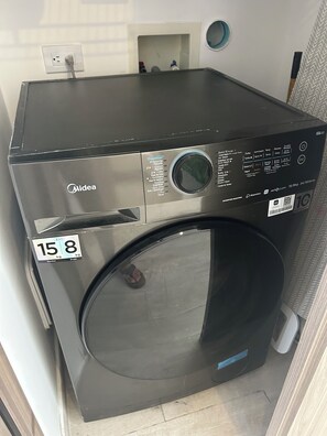 Eine schwarze Midea Waschmaschine mit einem Aufkleber, der eine Kapazität von 15 kg angibt, steht in einem Raum mit einer weißen Wand und einem kleinen Regal.
