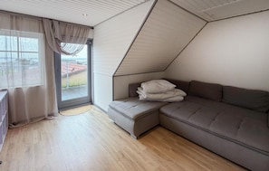 A room with a sloped ceiling, a grey sofa, a window with curtains, and a view of a building outside. - 3 bedroom beautiful home in Broager (Broager)