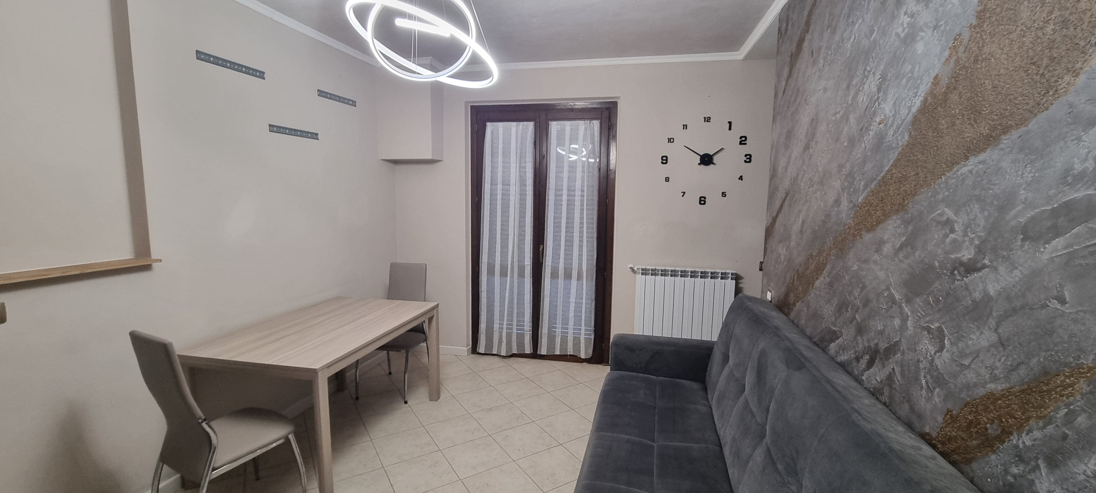 A room with a dining table, chairs, a sofa, a clock, and a radiator.