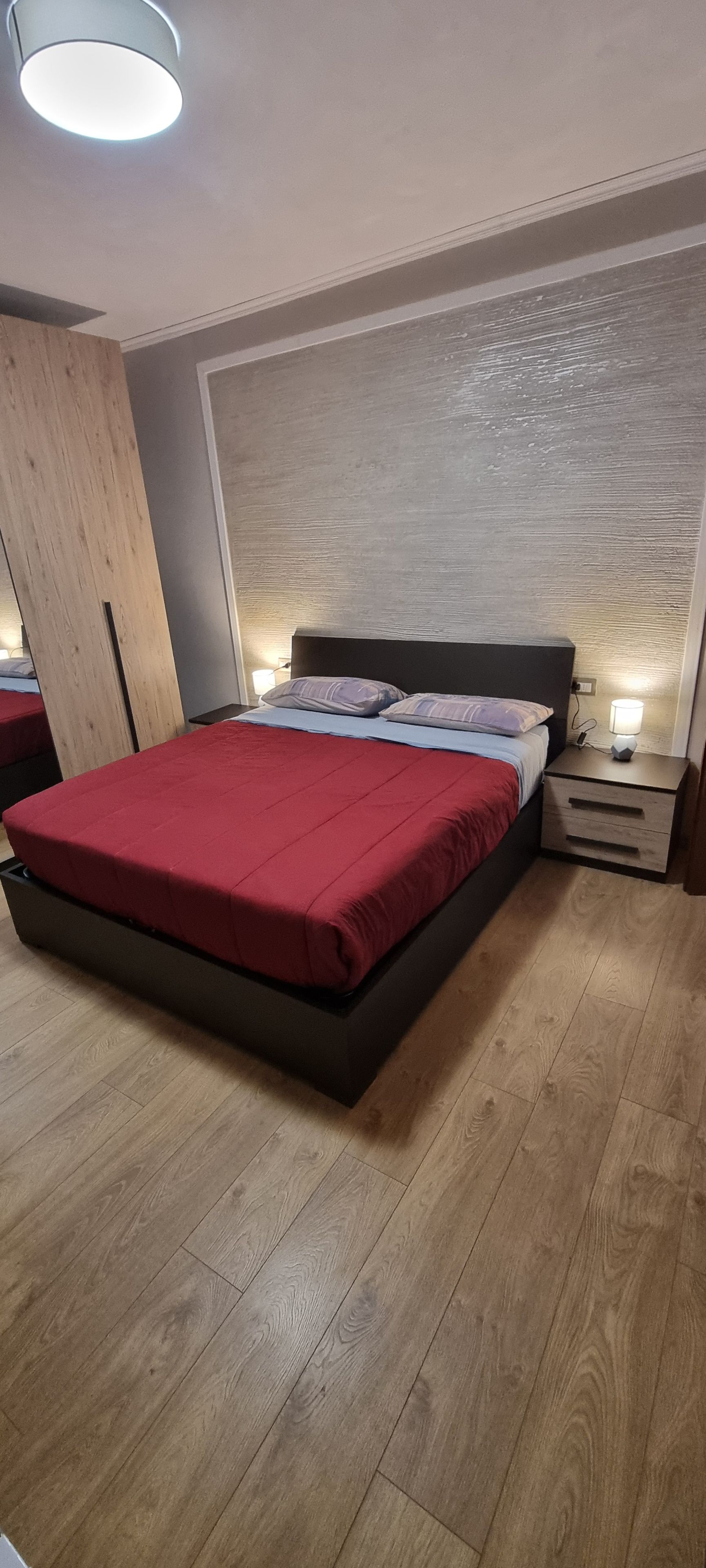 A modern bedroom with a large bed, wooden flooring, and a nightstand with a lamp.