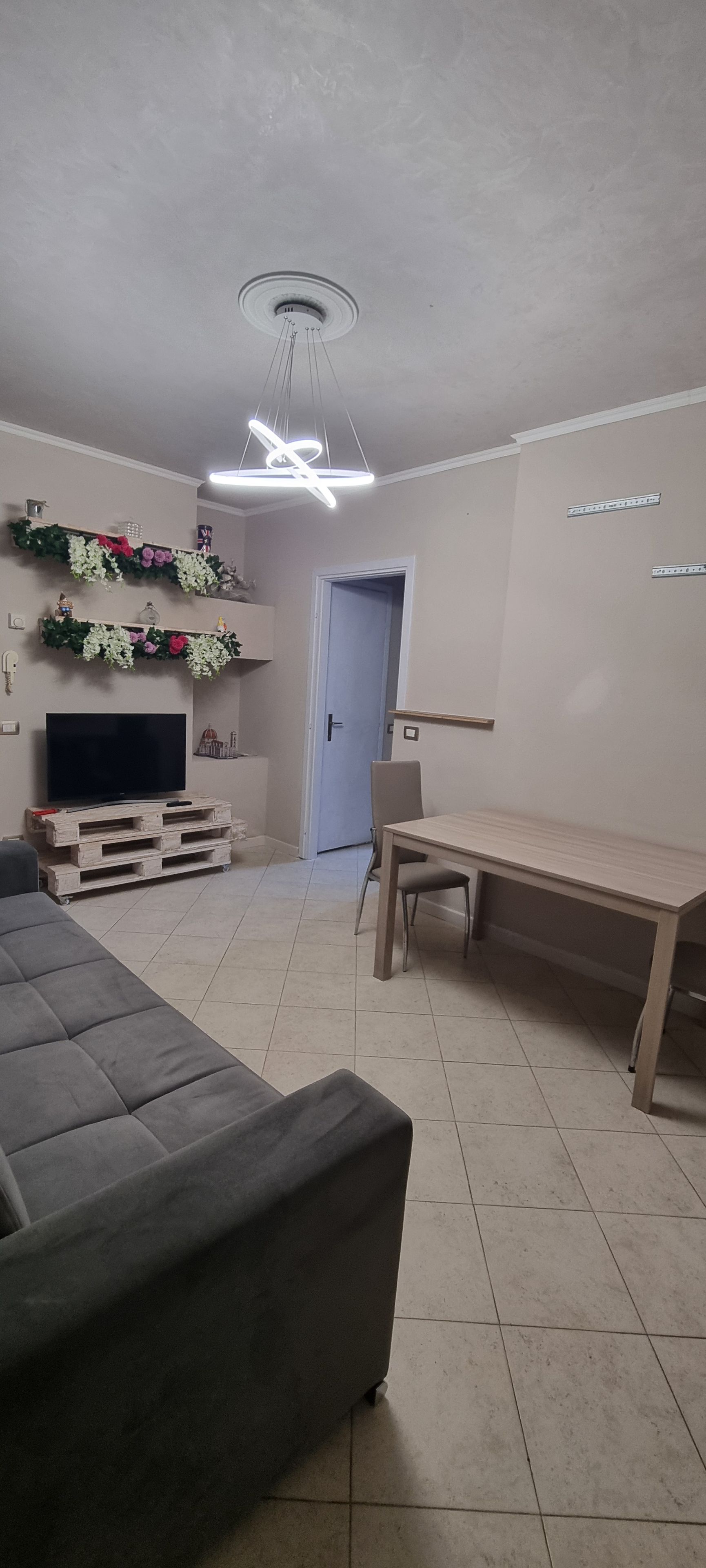 Una sala de estar con un sofá, un televisor sobre un mueble de madera, una mesa de comedor con sillas, una lámpara de techo y una puerta al fondo.