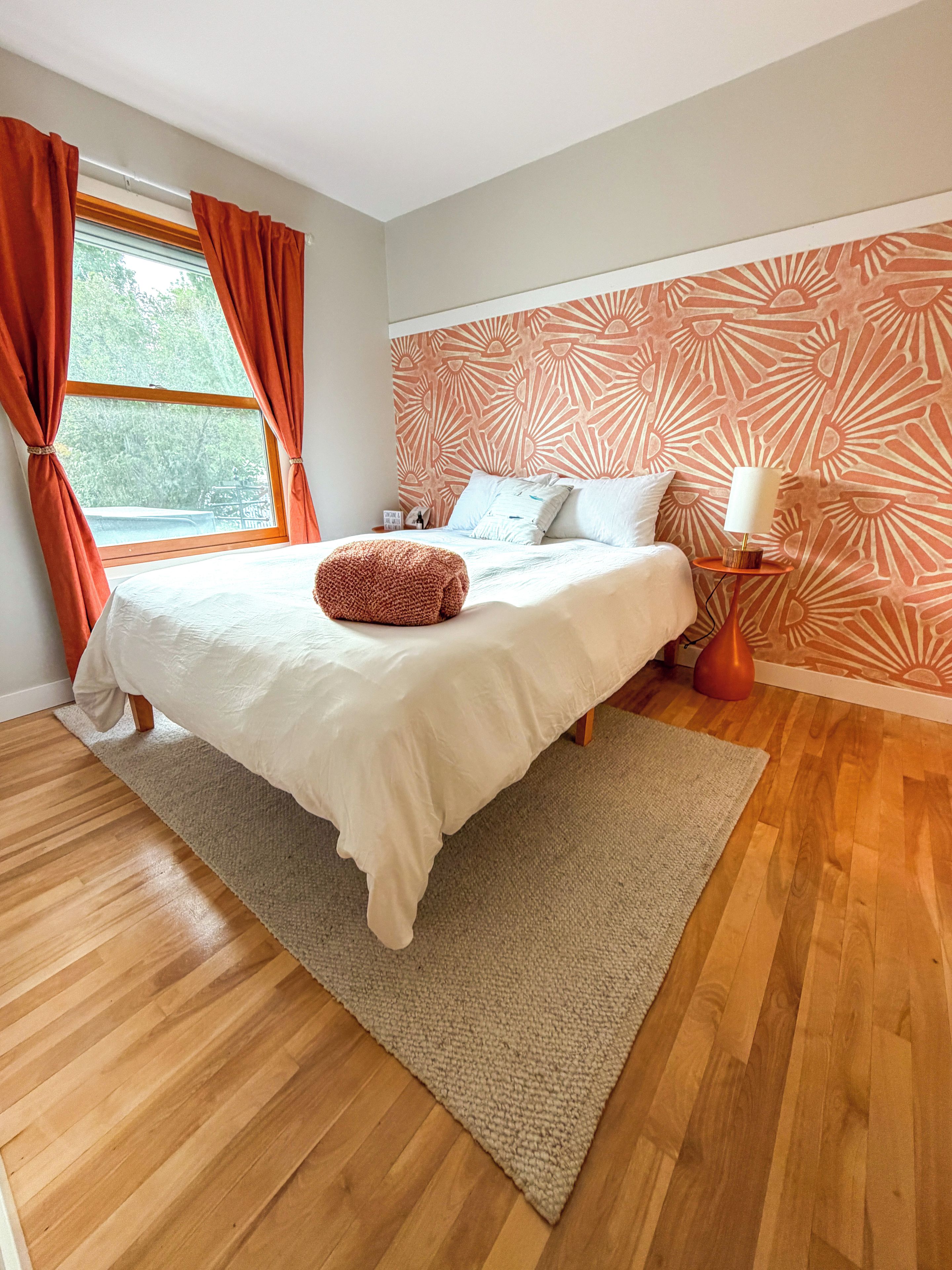 Un dormitorio con una cama grande, piso de madera, ventana con cortinas y pared estampada.