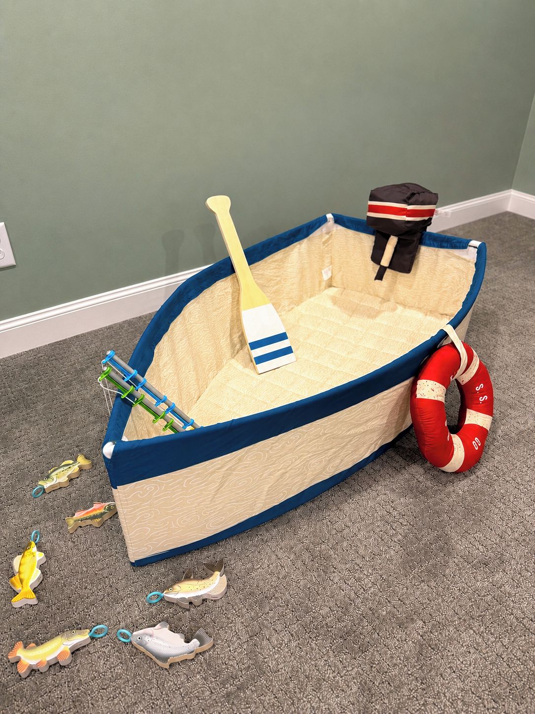 Ein Spielzeugboot mit einem Paddel, einer Rettungsboje und Spielzeugfischen auf einem Teppichboden.