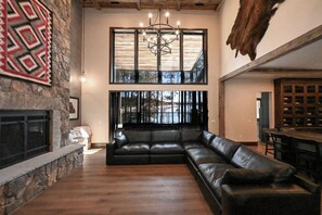 A spacious living area with a stone fireplace, a black leather sofa, and a large window offering a view of the outdoors. - Misty Mountain Lakefront Lodge with Dock and Hot Tub (Island Park)
