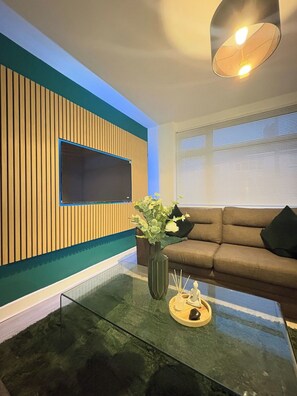 A modern living room with a flat-screen TV mounted on a wooden panel wall, a glass coffee table with a vase and candle, and a brown sofa with cushions. - Contractor Stay, Sleeps 5, Parking, WiFi, Halton (Halton)