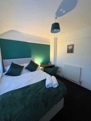 A neatly made bed with green bedding and pillows, a bedside table with a lamp, a radiator, and a wall-mounted picture. - Contractor Stay, Sleeps 5, Parking, WiFi, Halton (Halton)