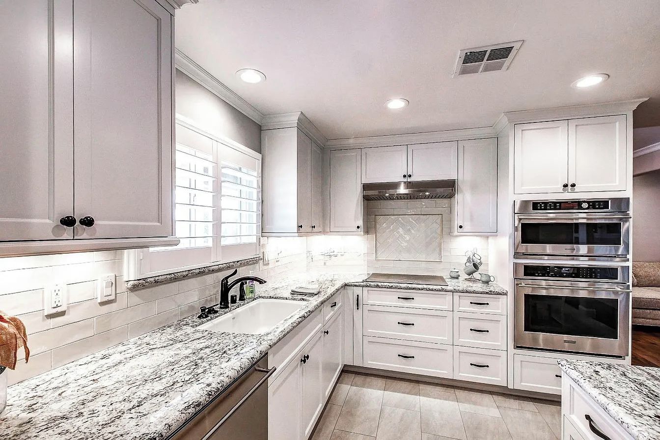 A modern kitchen with white cabinetry, granite countertops, and stainless steel appliances including a double oven and a range hood.