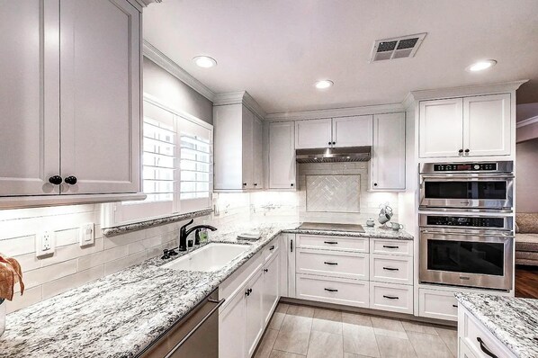 A modern kitchen with white cabinetry, granite countertops, and stainless steel appliances including a double oven and a range hood.