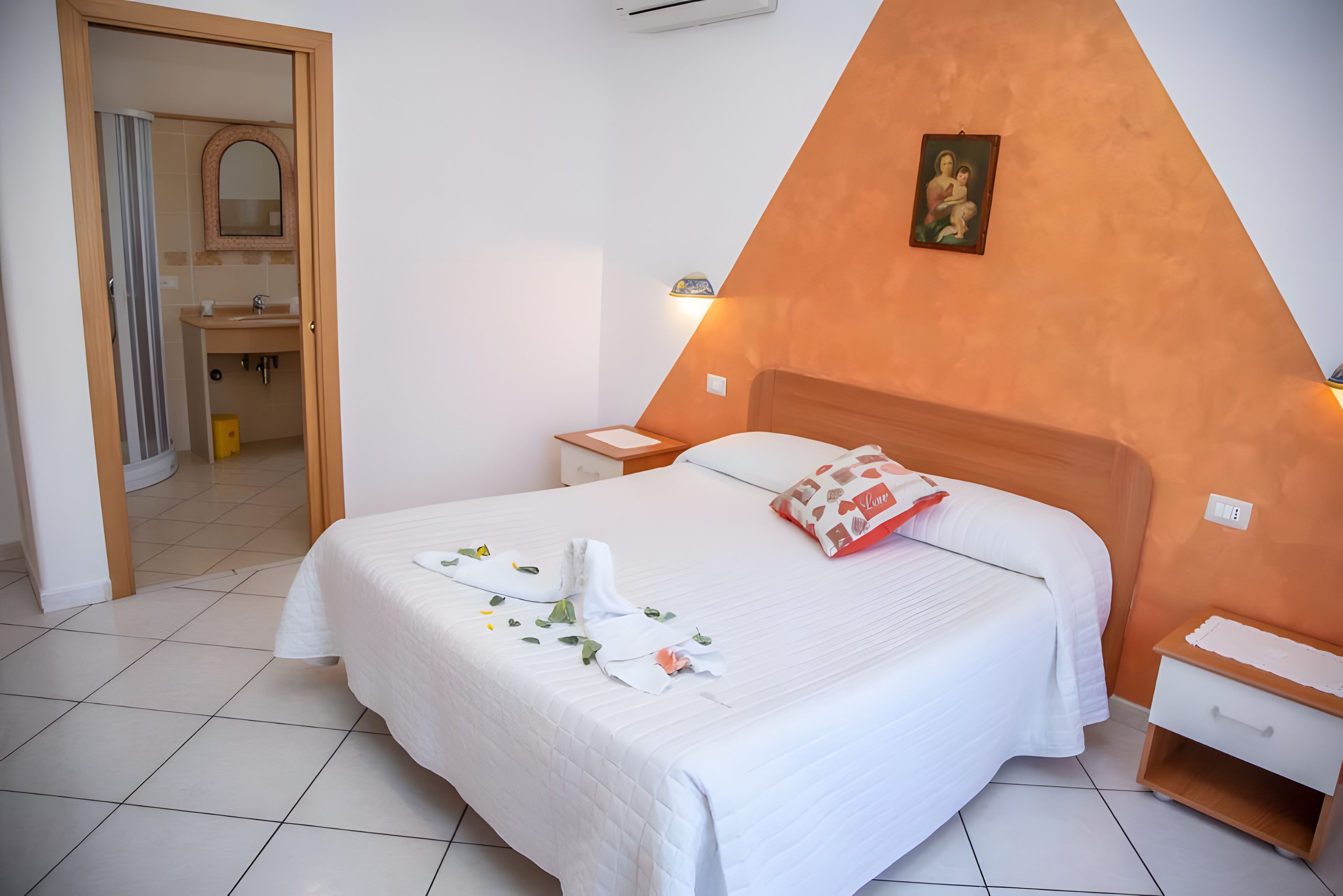 A hotel room with a bed, bedside tables, a nightstand, a mirror, and a bathroom visible through an open door.