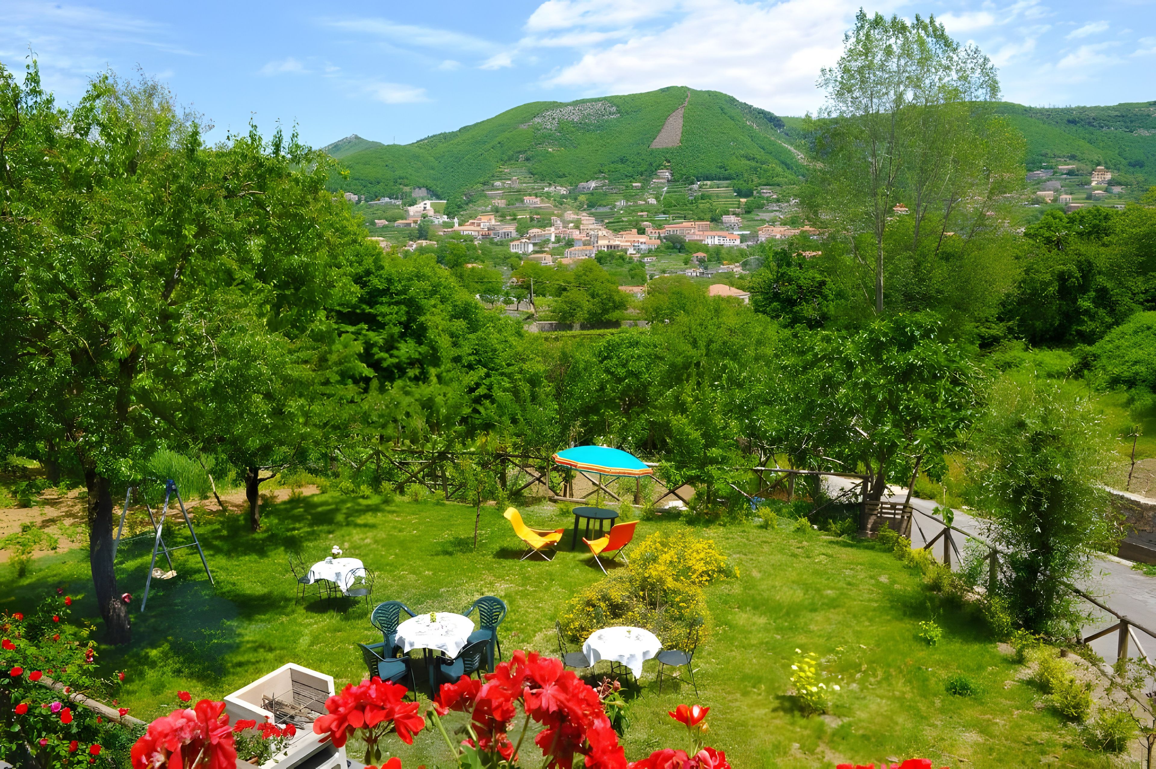 Un jardin luxuriant, avec une grande variété de plantes et de fleurs, un espace aménagé pour pique-niquer avec une table et des chaises, et une vue sur une ville et des collines situées au loin.