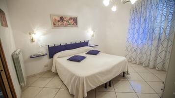 Un dormitorio con cama, mesita de noche, luz de pared, radiador, ventana con cortinas y un cuadro en la pared.