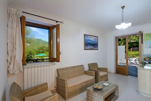 Une pièce donnant sur un paysage verdoyant grâce à une fenêtre ouverte, avec un canapé, un fauteuil, une table sur laquelle se trouve un bol, et une porte menant à l’extérieur.