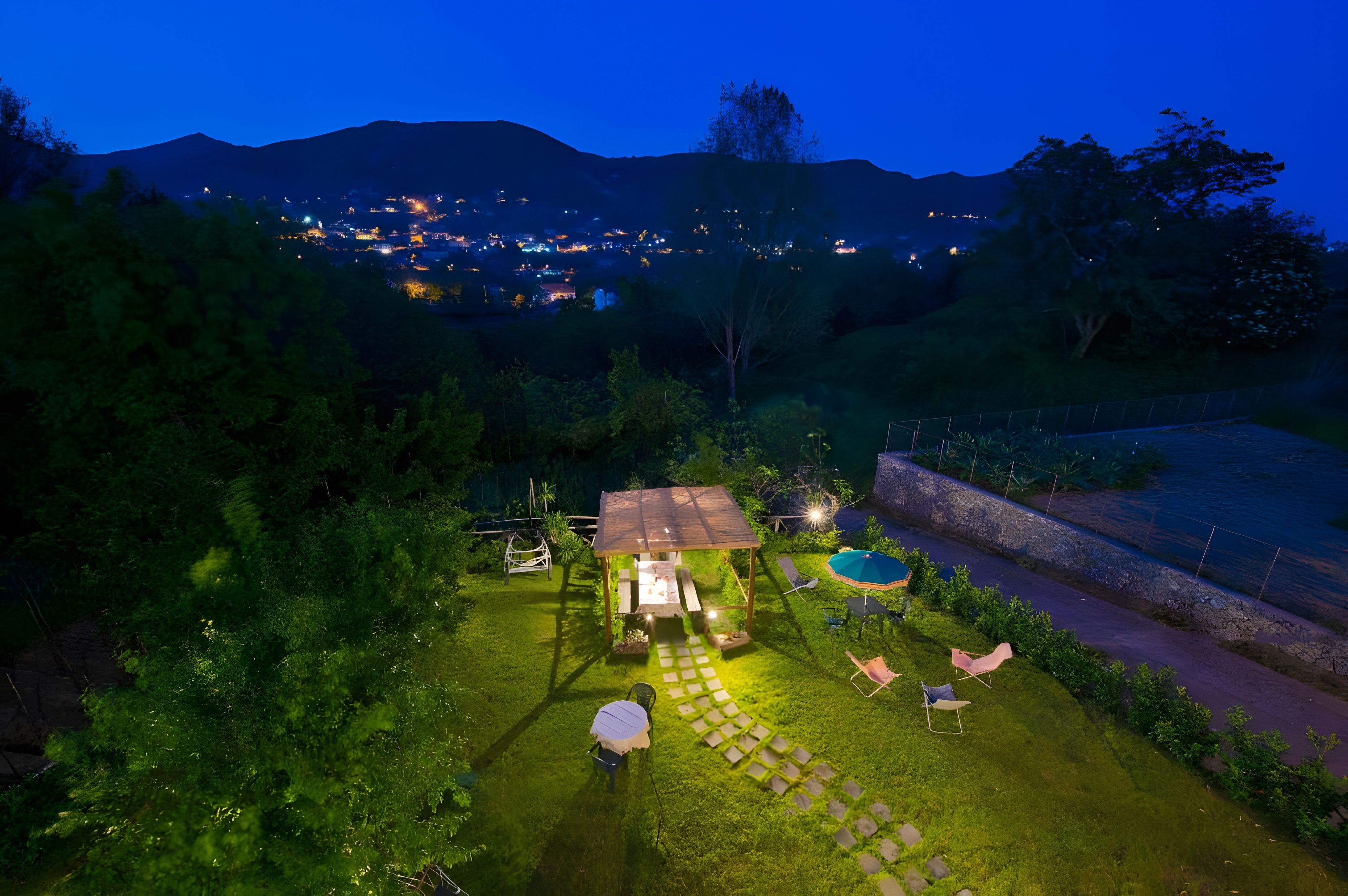 Un'area esterna ben illuminata con una zona salotto coperta, una piscina e vista su una città in lontananza di notte.