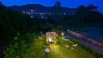 Un'area esterna ben illuminata con una zona salotto coperta, una piscina e vista su una città in lontananza di notte.