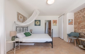 Ein Schlafzimmer mit einem Bett, einem Nachttisch, einer Lampe, einem Sessel und einem Tisch. Durch eine offene Tür ist eine Dusche erkennbar.