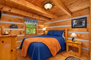 Ein Schlafzimmer in einer Blockhütte mit einem Bett, zwei Nachttischen, einem Fenster mit Blick auf Bäume und einer Wandleuchte.