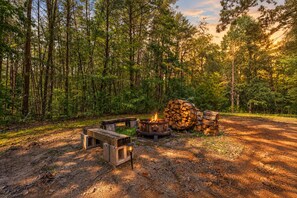 Ein Campingplatz mit Feuerstelle, Bänken und einem Holzstoß in bewaldeter Landschaft.