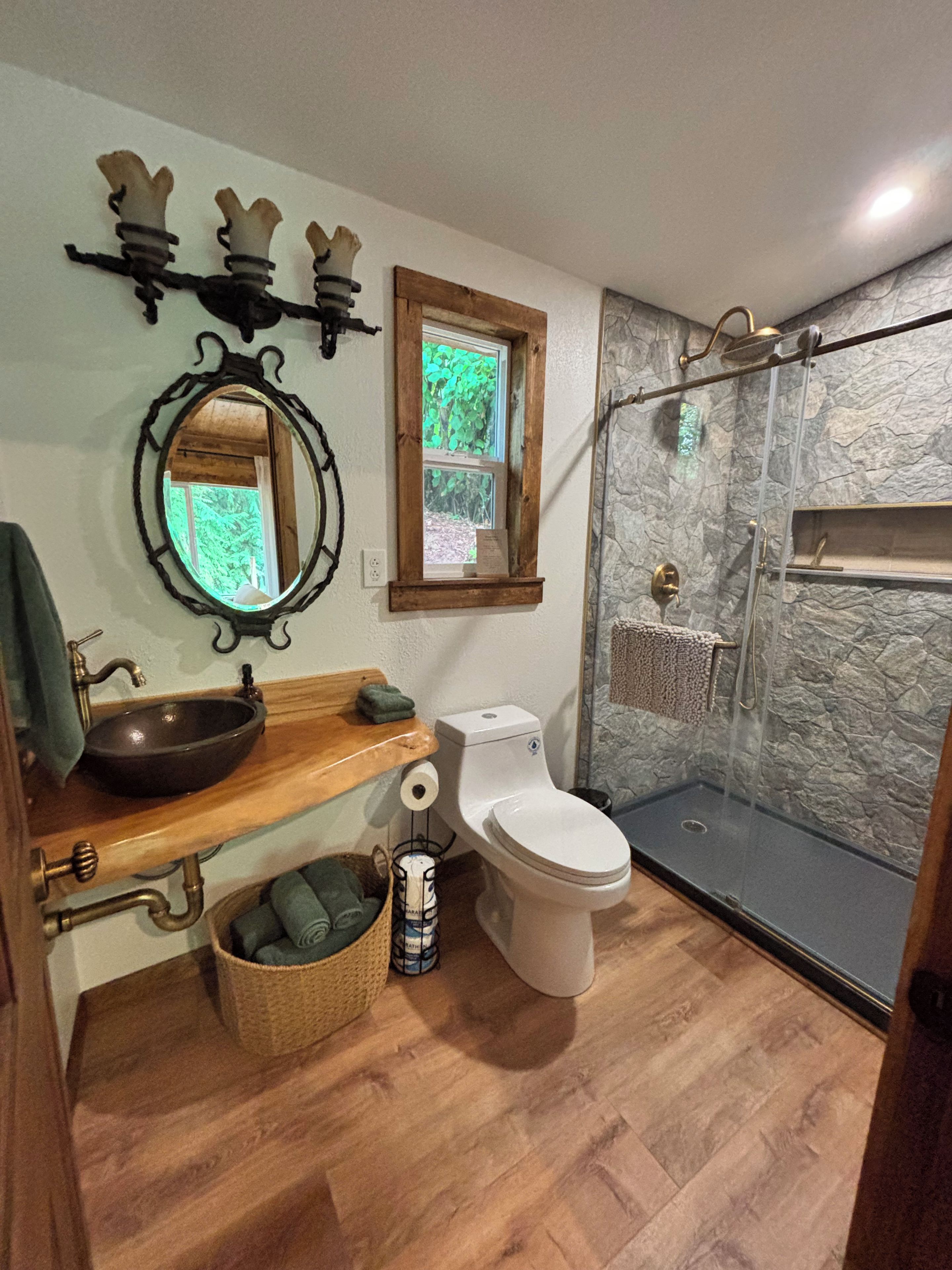 A bathroom with a wooden vanity, a round mirror, a toilet, and a glass-enclosed shower.
