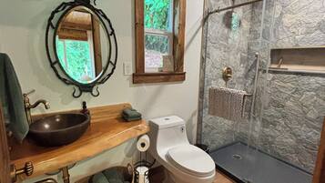 A bathroom with a wooden vanity, a round mirror, a toilet, and a glass-enclosed shower.