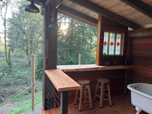 A rustic cabin with a wooden bench, two stools, a vintage bathtub, and a window with stained glass.