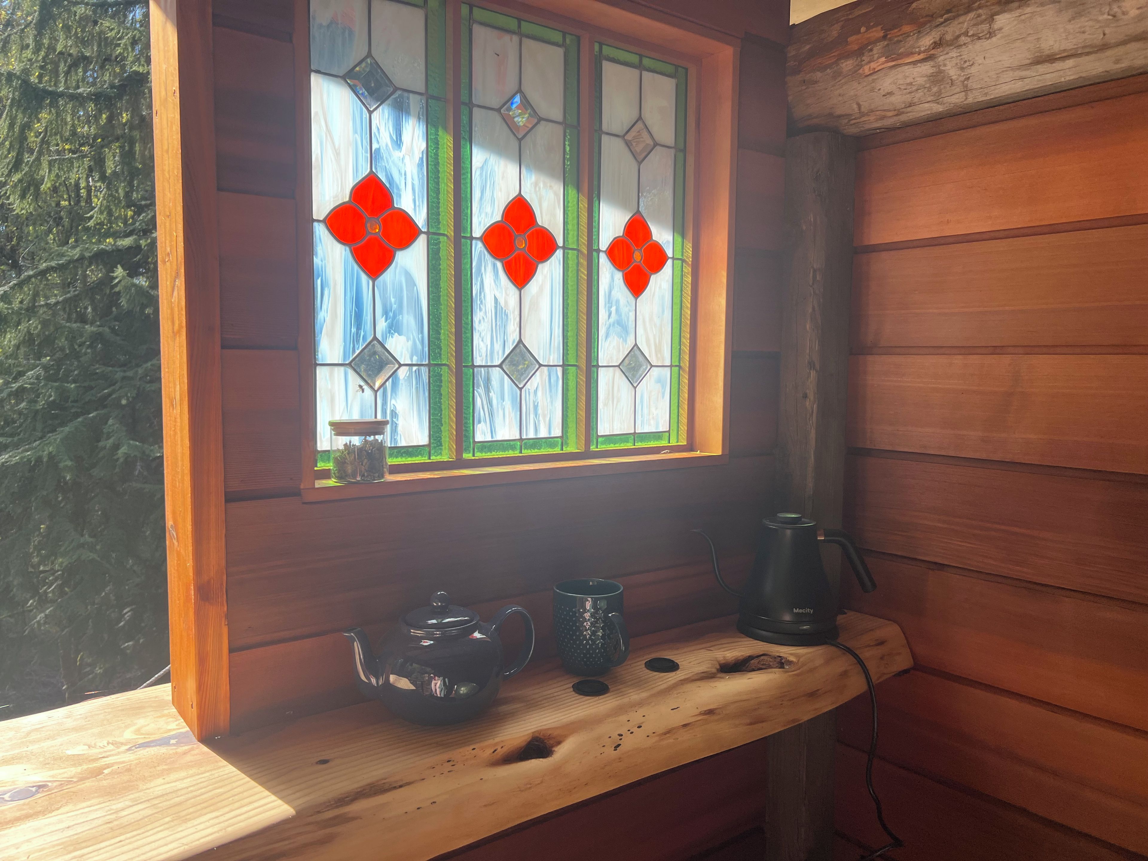 A wooden cabin interior with a window featuring stained glass with red diamond shapes, a wooden shelf with a teapot, a mug, and a kettle.