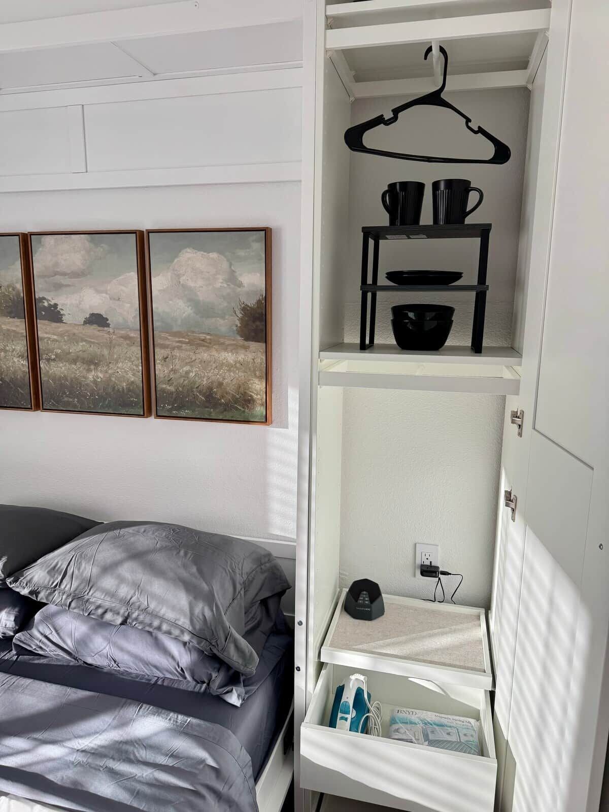 Une chambre à coucher avec un lit, une armoire blanche avec une tringle pour suspendre les vêtements et une étagère contenant des ustensiles de cuisine.