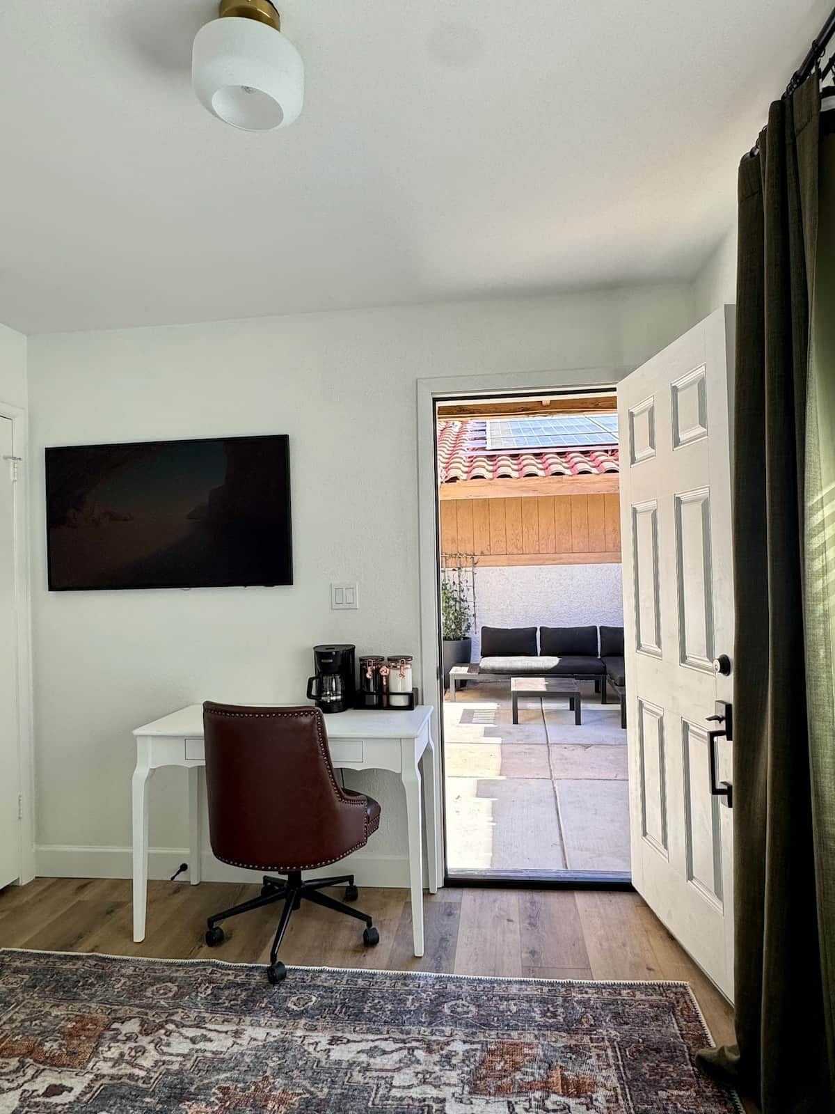 Una habitación con un escritorio blanco, una silla de oficina marrón, un tapete estampado, un cuadro en la pared y vistas a un patio exterior con un sofá y una mesa.
