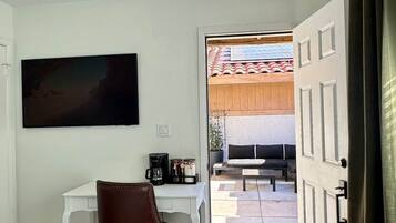Una habitación con un escritorio blanco, una silla de oficina marrón, un tapete estampado, un cuadro en la pared y vistas a un patio exterior con un sofá y una mesa.