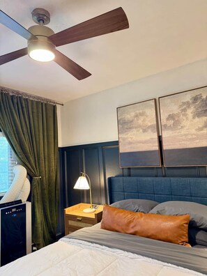Hotelkamer met een bed, een plafondventilator, een nachtkastje met een lamp, een raam met gordijnen en twee aan de muur hangende schilderijen.