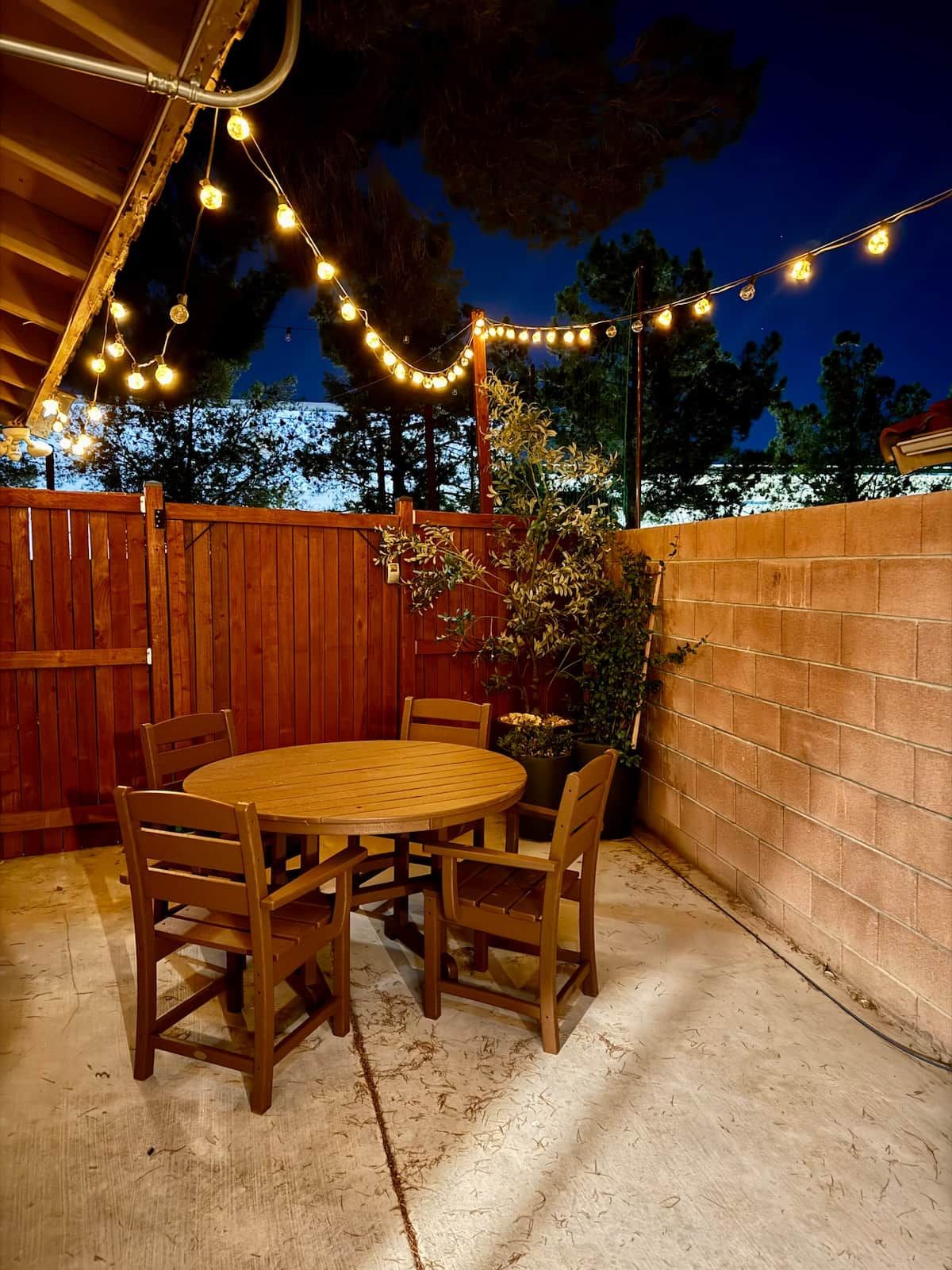 Un patio al aire libre con una mesa y sillas de madera, rodeado por una valla de madera y un muro de ladrillo.