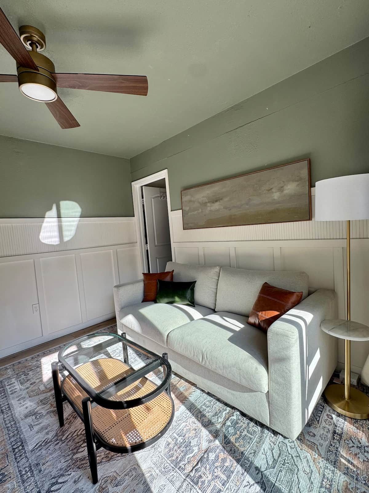 Un salon avec un canapé, une table basse en verre, un ventilateur de plafond, un tableau au mur et une lampe.