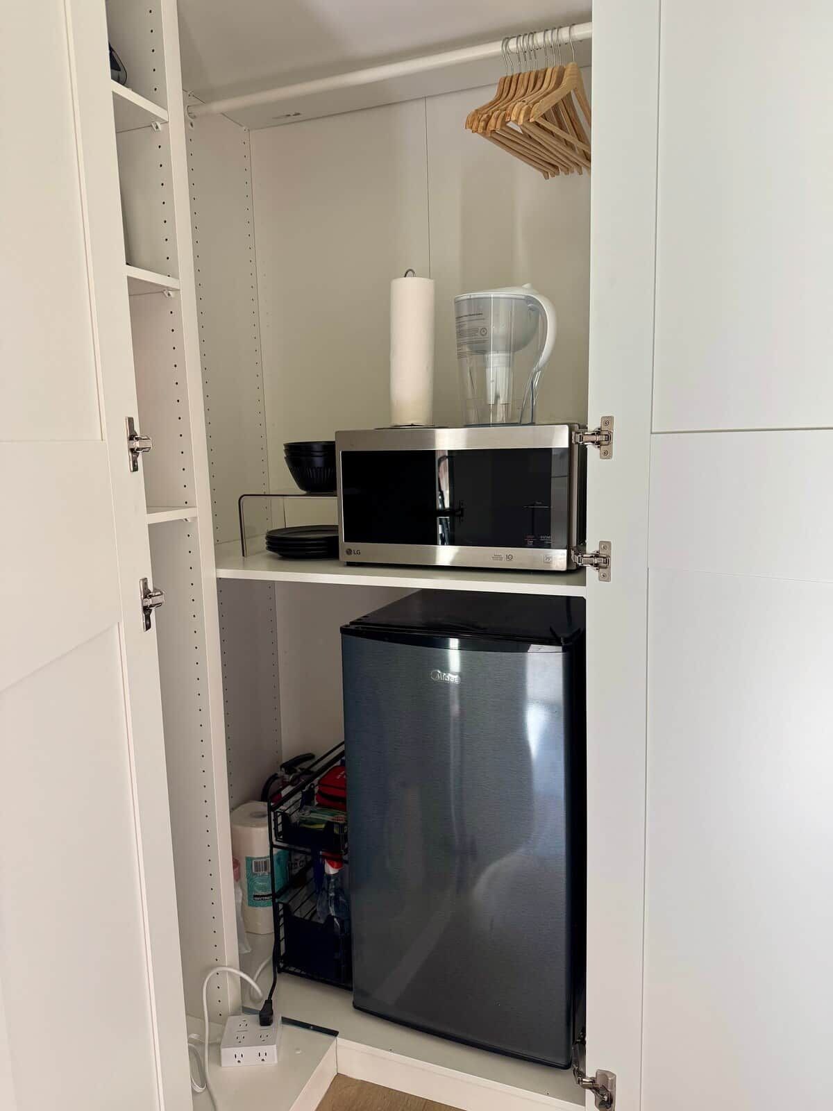 Una kitchenette compacta con microondas, un refrigerador pequeño y un estante de madera colgante.