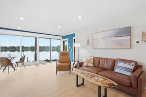 A modern living room with a view of a lake, a brown leather sofa, a wooden coffee table, and a dining area with a table and chairs.