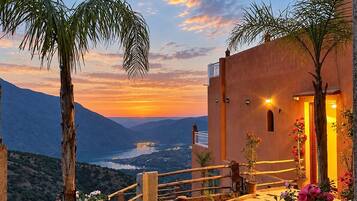 A sunset view from a terrace with a stone pathway, palm trees, and a building with a balcony.
