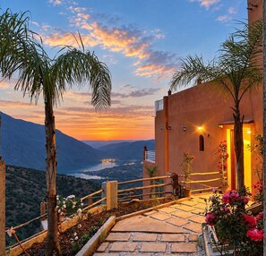 A sunset view from a terrace with a stone pathway, palm trees, and a building with a balcony. - Riad Ouirgane Panorama (Ouirgane)