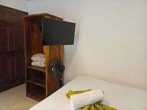 A room with a wooden wardrobe, a flat-screen TV mounted on it, a standing fan, and a bed with a white mattress and a green towel. - Apartamentos y Villas D Angelo - Superior Apartment 101 (Manzanillo)
