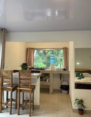 A compact kitchen with a dining area, a water dispenser, and a bed with a view of greenery. - Apartamentos y Villas D Angelo - Superior Apartment 101 (Manzanillo)