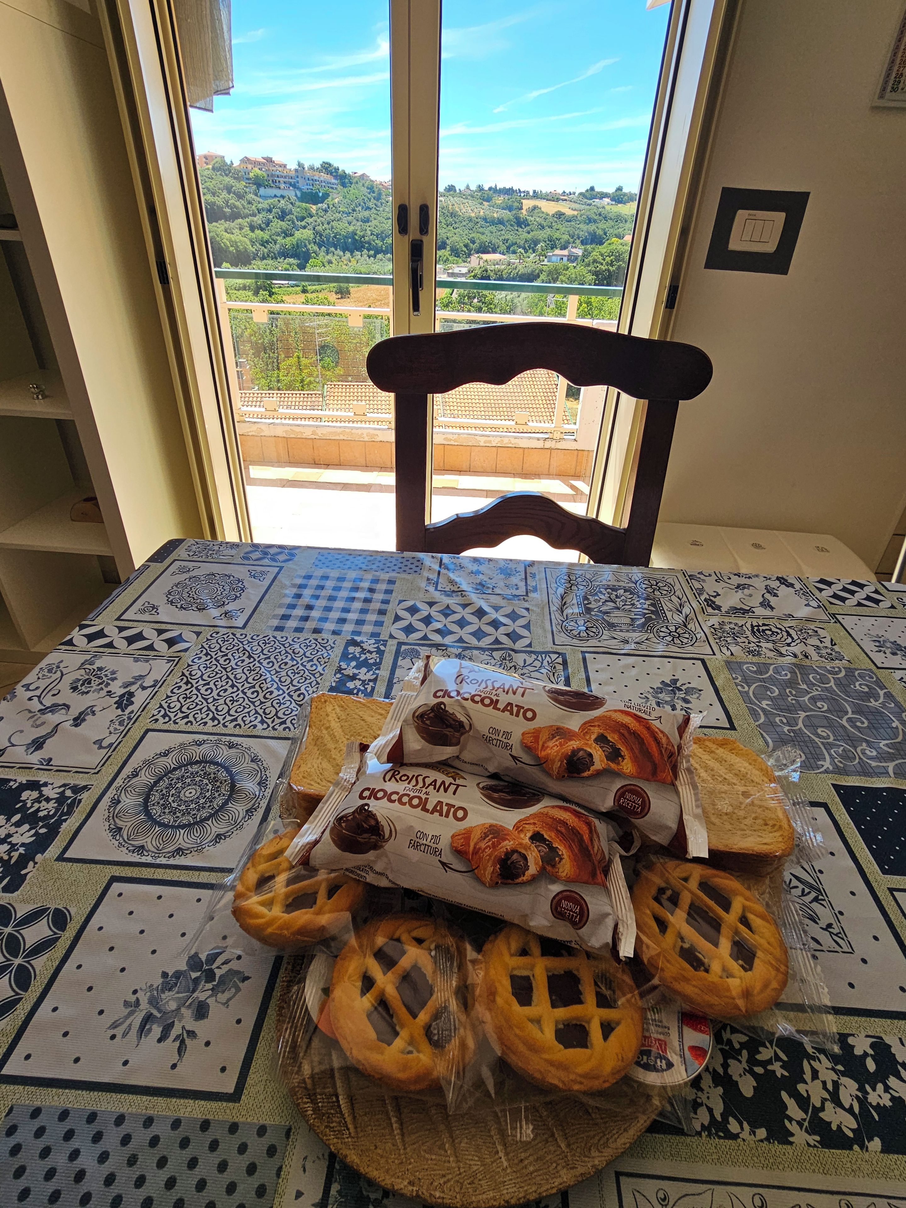 A table with a patterned tablecloth, a chair, and a view of a green landscape through a window.