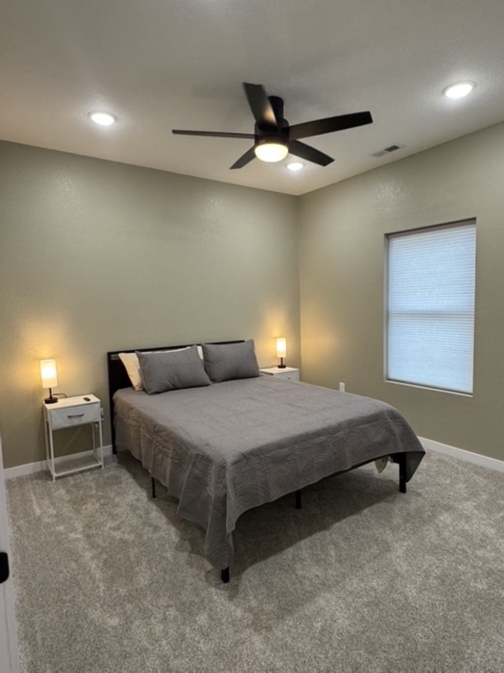 A bedroom with a bed, two bedside tables with lamps, a ceiling fan, and a window with blinds.