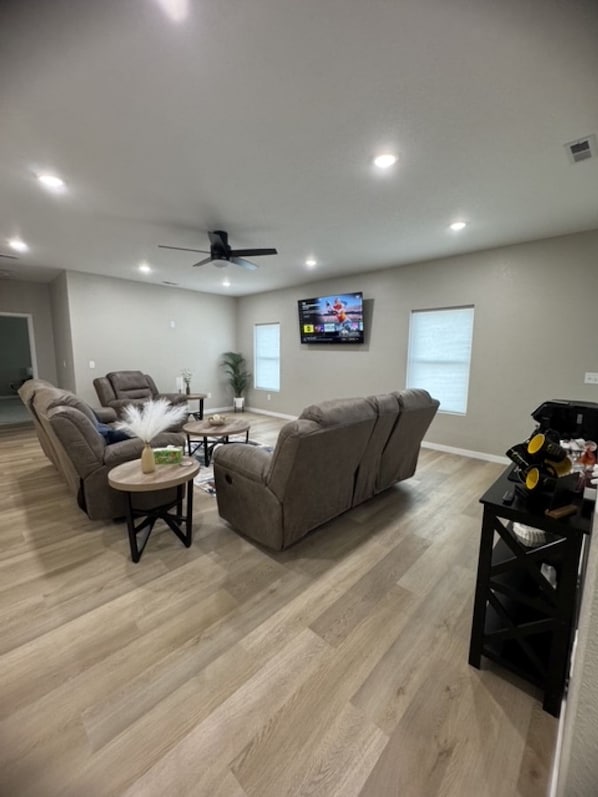 A spacious living room with a ceiling fan, a flat-screen TV mounted on the wall, and a seating area with a sofa and chairs.