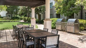 Un patio exterior con mesas y sillas, una parrilla y una zona cubierta con columnas.